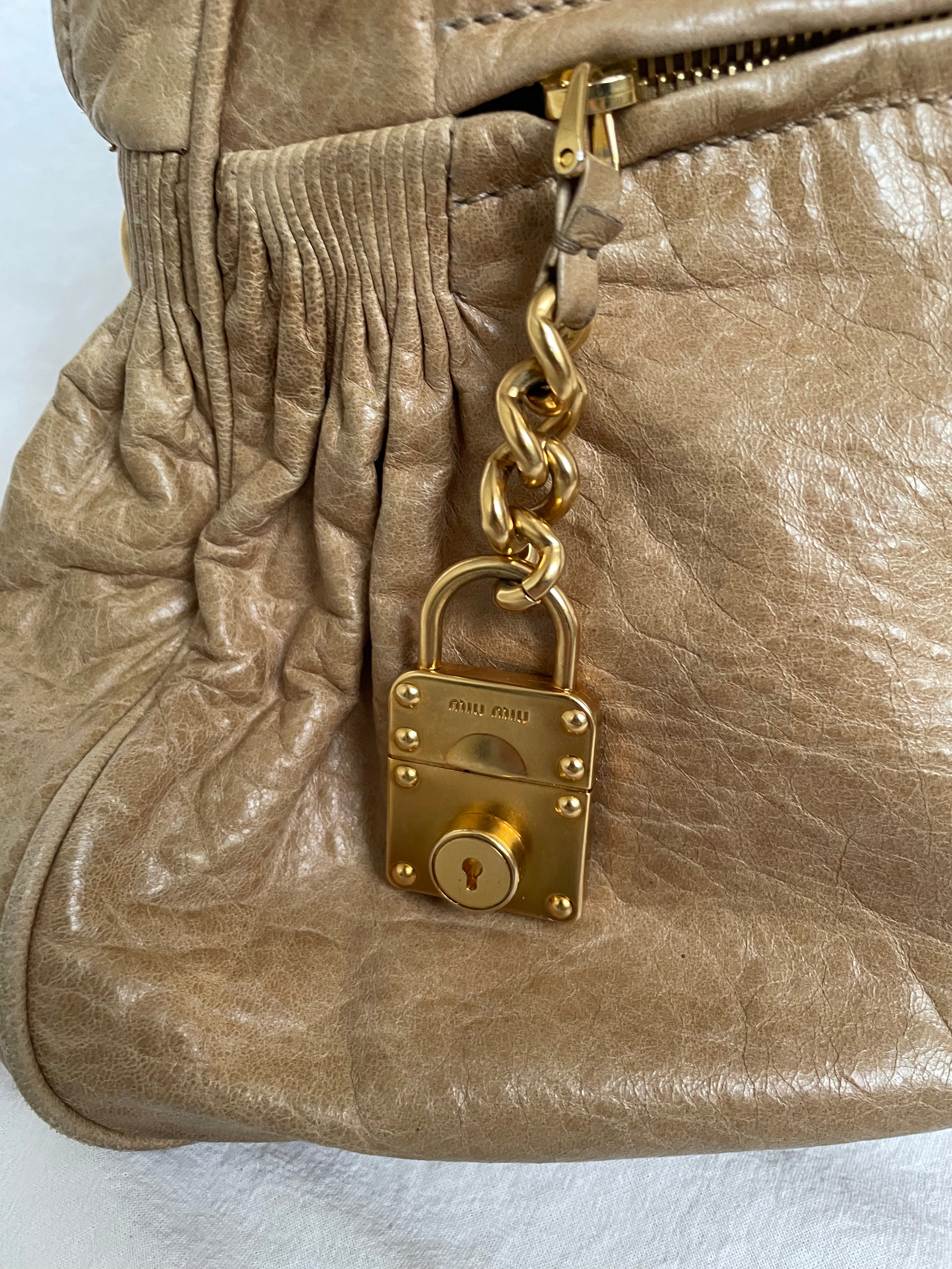 Miu Miu leather handbag. Crafted from matelassé calf leather, this handbag features luxurious lambskin and gold-tone hardware. It includes a front zip pocket, a top zip closure, and an interior slip pocket. The bag has a gold padlock charm.&nbsp;