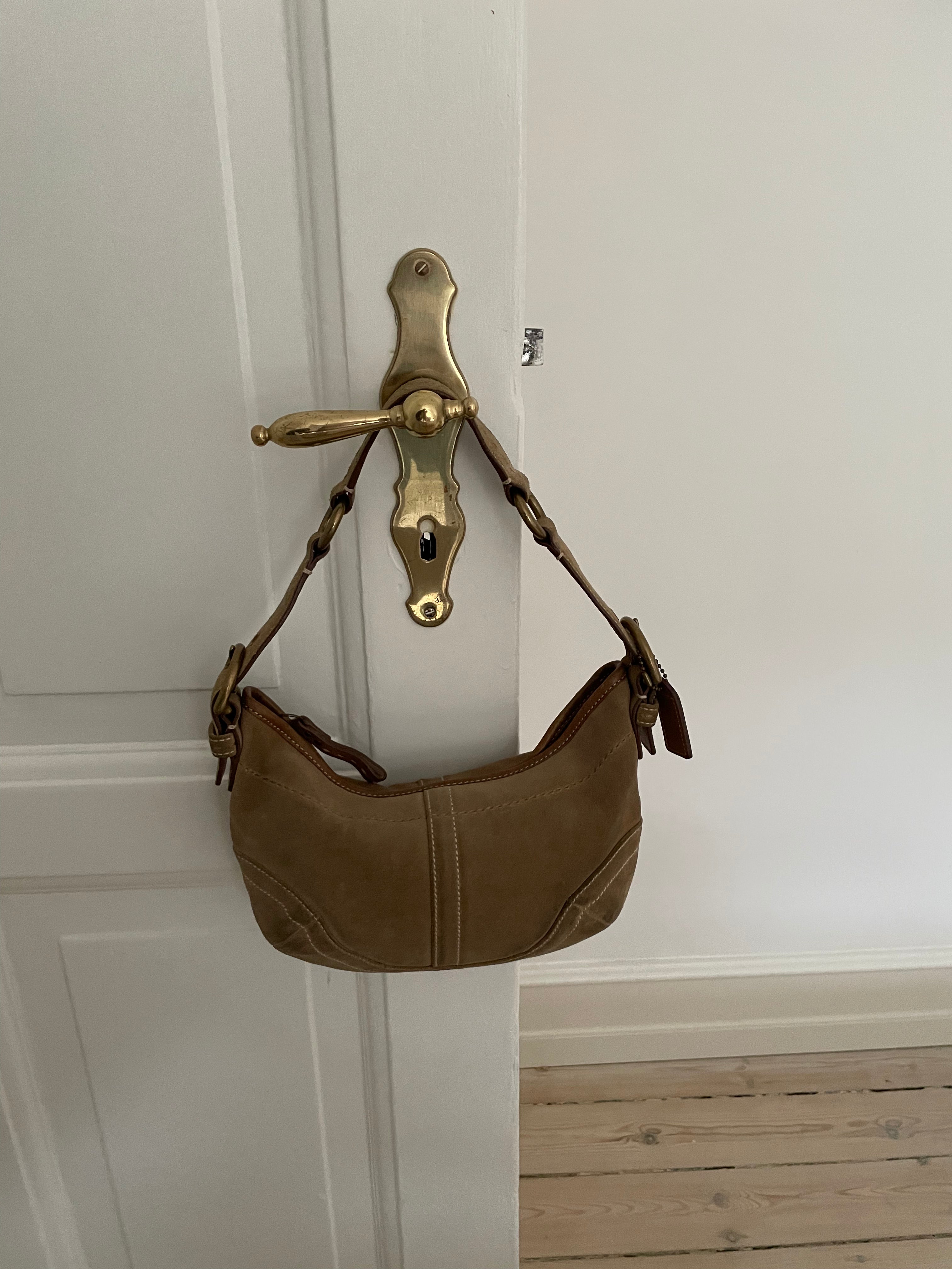 This beautiful Coach shoulder bag, crafted from light brown suede, is in good condition. It features unique white stitching, an adjustable leather strap, and offers an interior zip pocket along with one open pocket.